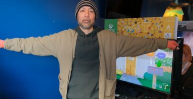 Keita Takahashi holding concept art for To a T, smiling with colorful game visuals in the background