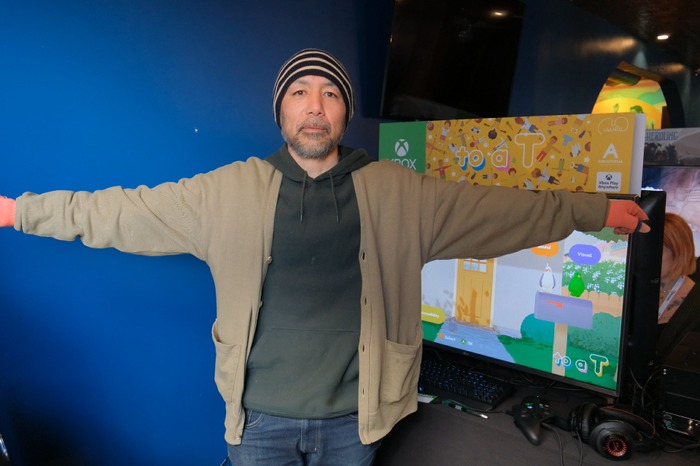Keita Takahashi holding concept art for To a T, smiling with colorful game visuals in the background